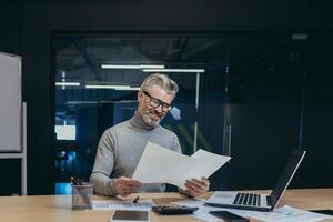 Happy and smiling mature businessman behind paper work, gray haired boss working with accounts and documents, man working in modern office, successful investor reviewing financial reports photo