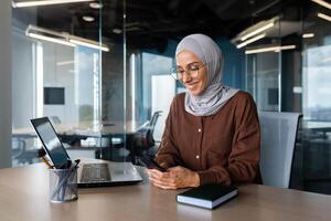 Young muslim business woman, freelancer in hijab working in office at desk and using phone smiling. Chats, types and reads messages. photo