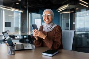 Smiling young Muslim woman in hijab working in office on laptop, using mobile phone, smiling at camera. photo