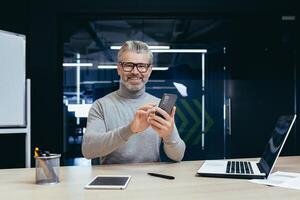 Portrait of successful mature investor inside office with work, gray-haired businessman smiling and looking at camera, man holding phone, typing message, using laptop at work. photo