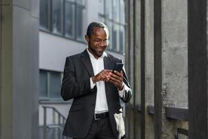 Happy successful African American businessman boss walking outside office building, manager holding smartphone reading message and smiling, using communication app. photo