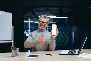 Portrait of mature businessman inside office, senior boss smiling looking at camera and showing white blank smartphone screen, man holding affirmative and recommending thumbs up working with laptop. photo