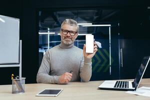 Successful businessman inside office showing phone with white screen free space to camera, mature boss working on laptop with documents, man smiling satisfied recommending showing banner. photo