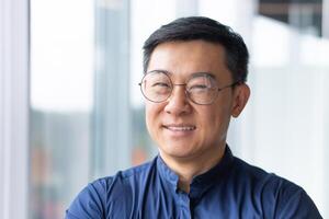 Close-up portrait of smiling asian man inside office, man smiling and looking at camera in glasses and shirt, successful engineer programmer engineer developer in development company at work. photo