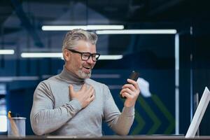 Mature businessman with beard using app on phone inside office, man received online message notification about good news, boss texting online at workplace. photo