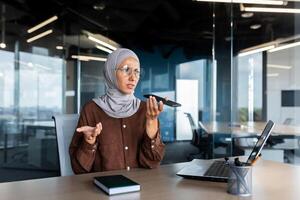 Worried young Muslim businesswoman in hijab sitting in office and talking on the phone through loudspeaker, recording voice message, solving questions, problems. photo