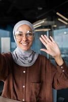 Close-up photo. Vertical photo. A young female blogger in a hijab talks to the camera, holds a phone in her hand. Films a blog, a podcast. He greets the camera with his hand, smiles. photo