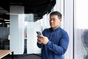 A serious and concentrated Asian man inside the office is using the phone, a businessman is standing by the window browsing the Internet using an application on a smartphone reading news and texting. photo