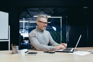 Cheerful and smiling mature businessman working inside office, senior gray haired man using laptop at workplace smiling satisfied with good results achievement. photo