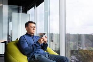 Serious thinking asian man reading news on phone, programmer using app on smartphone testing new program, businessman worker sitting on soft chair near window in modern office interior. photo