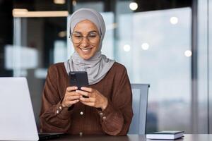 Close up of muslim successful businesswoman in hijab using phone inside modern office, workplace woman typing message and typing on smartphone screen smiling. photo