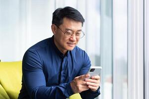 A young man, an Asian student, sits on an ottoman by the window in an office, co-working space, campus. He holds the phone in his hands, dials a message, reads the news, checks the mail, smiles. photo
