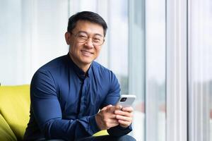 Portrait of a successful Asian man inside the office, a man sits near window smiling and looking at the camera, a businessman holds a phone in his hands, types a message and browses the Internet. photo