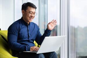 call, successful asian businessman near window in office talking remotely, online meeting with colleagues, programmer waving at laptop camera greeting gesture. photo