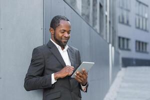 Photo from below. An African American businessman is standing outside an office center in a suit and using a tablet. Dials, communicates, smiles.