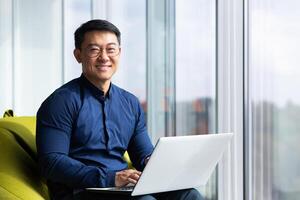 Portrait of successful asian businessman inside office, man smiling and looking at camera, using laptop at work near window, boss is satisfied with achievement results. photo