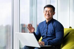 Portrait of successful smiling asian man inside office, man smiling and looking at camera near window, businessman working sitting with computer, waving hand in greeting gesture. photo