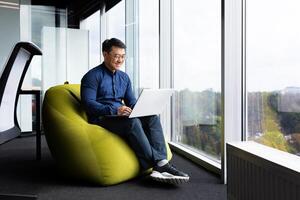 Successful mature asian working inside office using laptop, man typing on keyboard and smiling, businessman in shirt and glasses satisfied with work and achievement results, programmer at work. photo