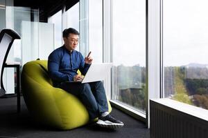 call, online remote conversation with clients and colleagues, successful asian man sitting in office near window on soft chair and talking remotely in meeting, man smiling using laptop at work. photo