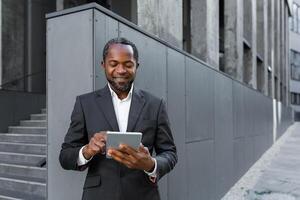 Successful african american man in business suit smiling and using tablet computer, businessman outside modern office building walking. photo