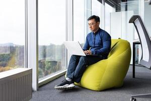 Serious and focused engineer developer programmer writing code using laptop at work to create programs and software, asian man inside modern office near window working. photo