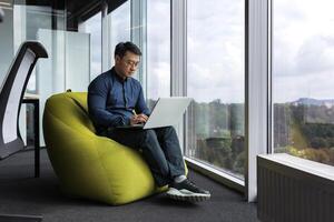 Asian young office worker sitting on soft ottoman chair, programmer using laptop for writing code and programming testing new program, man sitting near window inside office. photo