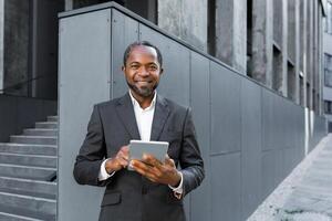 Portrait of a successful African American boss, a businessman in a suit smiling and looking at the camera, a man holding a tablet computer in his hands, a successful investor browsing the Internet. photo