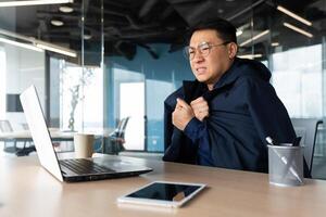 There is no heating in the office and the heater is broken, the businessman is freezing at the workplace, the man is sitting at the table inside the building using a laptop at work. photo