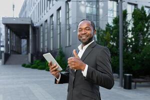 Portrait of a smiling and successful black senior businessman standing near an office center and working on a tablet. Looks at the camera and points a super finger. photo