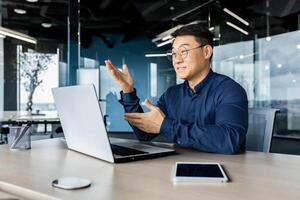 Successful Asian businessman talking on call using laptop for remote communication with colleagues, boss in shirt smiling and gesturing with hands, online meeting with partners. photo