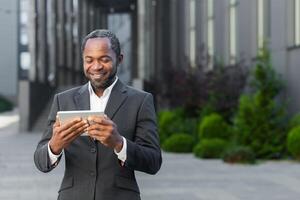 African american man in business suit outside office building watching online with using app on tablet computer, businessman smiling and happy. photo