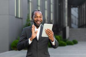An African American businessman in a suit stands outside an office center and talks on a call. He holds a tablet in his hand. He waves his hand at the camera, says hello, smiles. photo