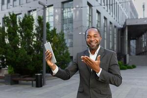 An African-American businessman is standing outside in a suit, holding a tablet and talking on a call. He looks at the camera, points to the gadget, smiles. photo
