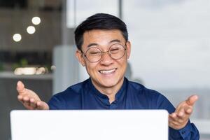 Close-up photo of successful Asian businessman talking remotely on call, male boss in glasses looking at laptop screen smiling and talking friendly, programmer working inside office.