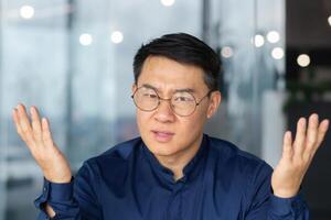 Angry boss close-up talking on call using laptop, man looking at web camera and arguing nervous businessman unhappy with online meeting with colleagues, asian man at work inside office angry. photo