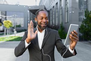 African american businessman in a suit and headphones standing outside an office center and talking on a call from a tablet. He waves his hand at the camera, says hello, smiles. photo