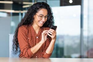 Beautiful woman in the office, happy and smiling latin american business woman uses internet phone close up, female worker reads message and browses internet pages inside office building. photo