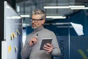 Mature businessman near a whiteboard with graphs analyzing and thinking about a strategy plan for the company's finances, the boss is holding a tablet computer and a marker for notes. photo