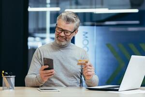 Cheerful and smiling gray-haired businessman inside a modern office makes purchases in an online store, the man holds a bank credit card and a smartphone in his hands, the buyer is satisfied choice. photo