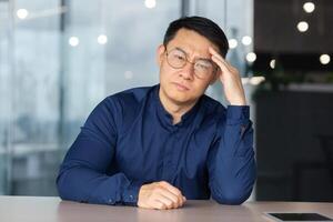 Portrait of overworked and tired office worker at workplace, asian man in glasses and shirt looking upset at camera, businessman not satisfied with work and own results achievement in depression. photo