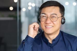 Closeup portrait man with a headset, Asian in glasses looking at the camera, employee is using headphones and microphone for a call, online customer service support tech, inside the office. photo
