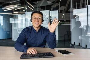 Webcam view, portrait of successful tech support and customer service worker, man smiling and looking at camera waving greeting hello, asian working inside modern office building. photo