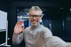 Mature businessman having fun talking on chat using app on smartphone, gray-haired man looking at phone camera and waving hands talking to colleagues while sitting at workplace inside office. photo