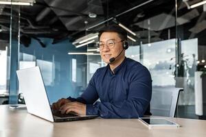 Happy and smiling worker with headset for call, Asian tech support man talking to online service users, businessman using laptop for online customer support. photo