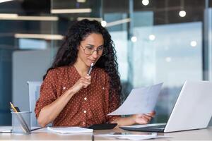 Beautiful hispanic woman in office doing paperwork, successful businesswoman financier working inside office sitting at table using laptop, at work with contracts and bills, thinking about decision. photo