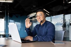 Successful smiling man working inside office using laptop and headset for call, asian cheerfully consulting tech support customer, businessman at work in shirt and glasses. photo