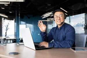 Man at work with headset phone and laptop successful asian smiling and looking at camera at workplace inside office, portrait of successful businessman support worker waving hand greeting gesture photo