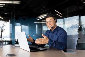 Asian with a headset is talking on call, man inside office smiling and gesturing joyfully in an online meeting, a businessman is telling his colleagues, a customer service tech support worker. photo