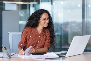 latin american businesswoman working inside office with documents and laptop, worker paperwork calculates financial indicators smiling and happy with success and results of achievement and work photo
