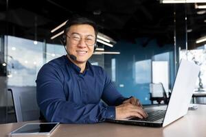 Portrait of successful office worker with headset for call, asian man smiling and looking at camera working inside office, man in shirt and glasses customer service support. photo